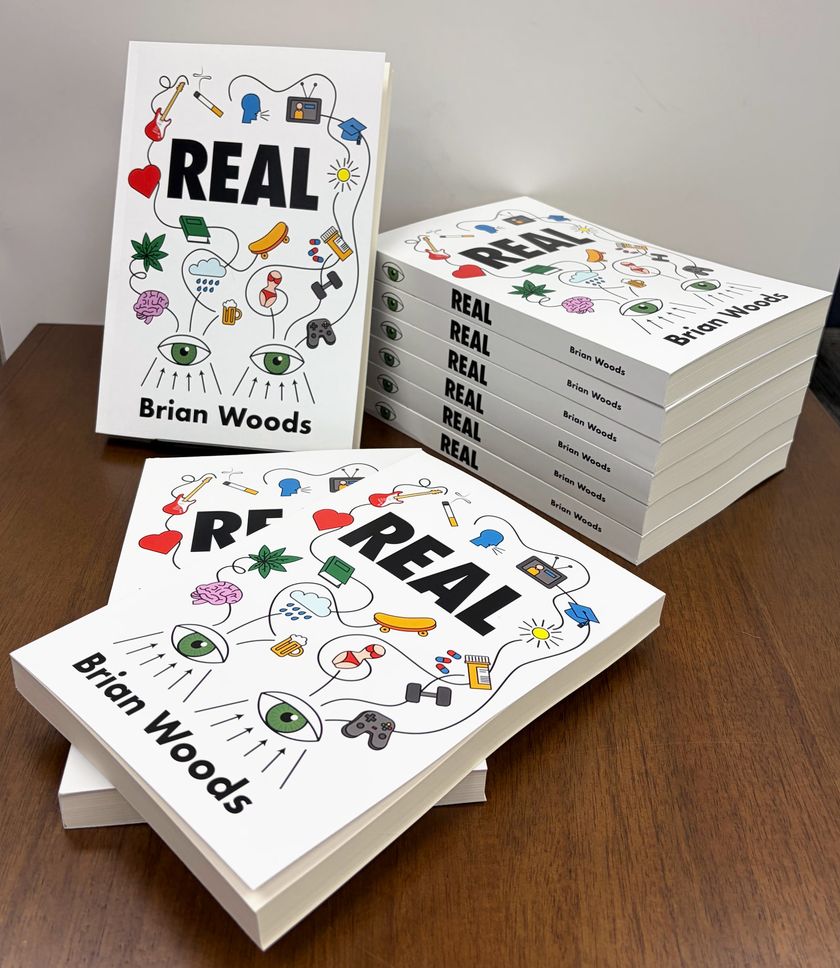 Nine paperback copies of Real, the novel by Brian Woods, on a wooden desk: one standing, six stacked, two lying flat.